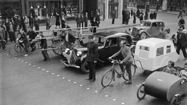 Dans une rue de Paris en 1940, un agent de police retient derrière une ligne de clous divers engins roulants