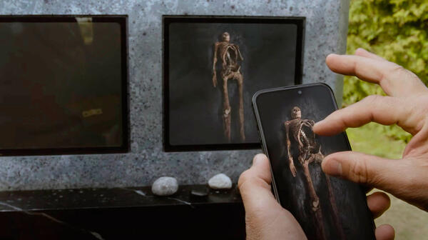 image du film où une main touche un téléphone qui montre l’intérieur de la tombe et on voit le squelette