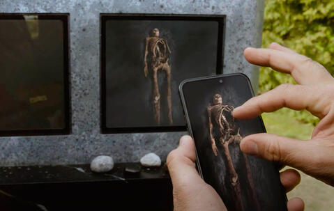 image du film où une main touche un téléphone qui montre l’intérieur de la tombe et on voit le squelette