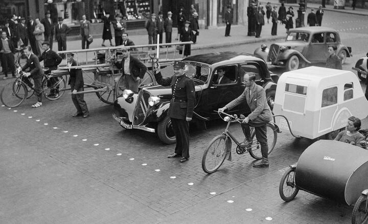 Dans une rue de Paris en 1940, un agent de police retient derrière une ligne de clous divers engins roulants