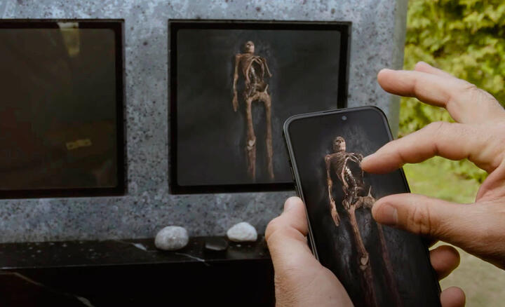 image du film où une main touche un téléphone qui montre l’intérieur de la tombe et on voit le squelette