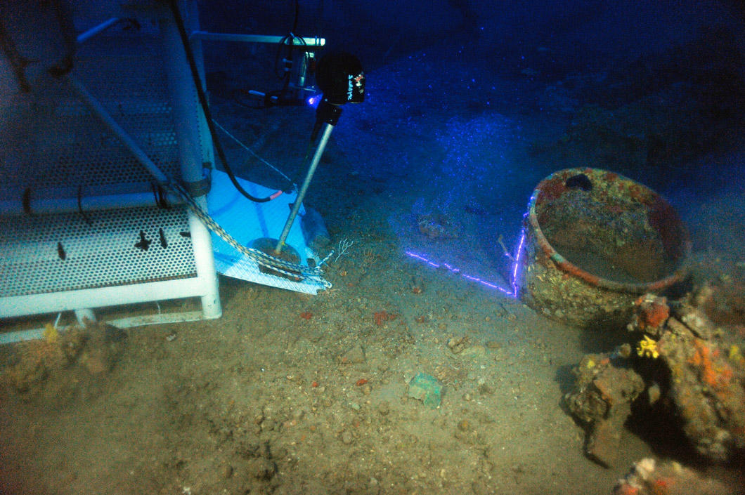 Sur le site d'une épave sous-marine, une machine scanne une jarre ensevelie avec un rayon lumineux violet.