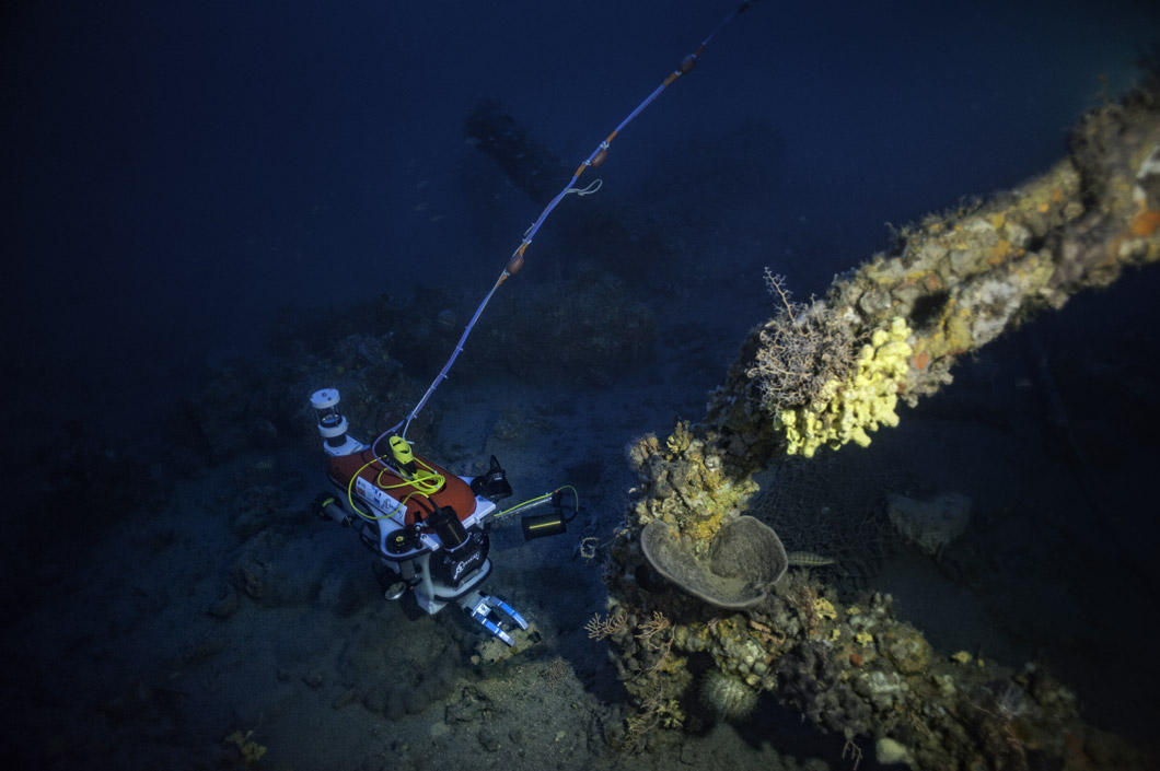 Le robot est au fond de l'eau. Au premier plan : l'ancre d'un bateau. On voit un câble qui part du robot.