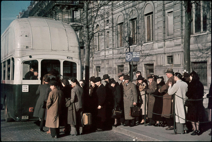 Photographie couleur restaurée montrant des gens qui font la queue pour monter dans un bus fonctionnant au gaz.