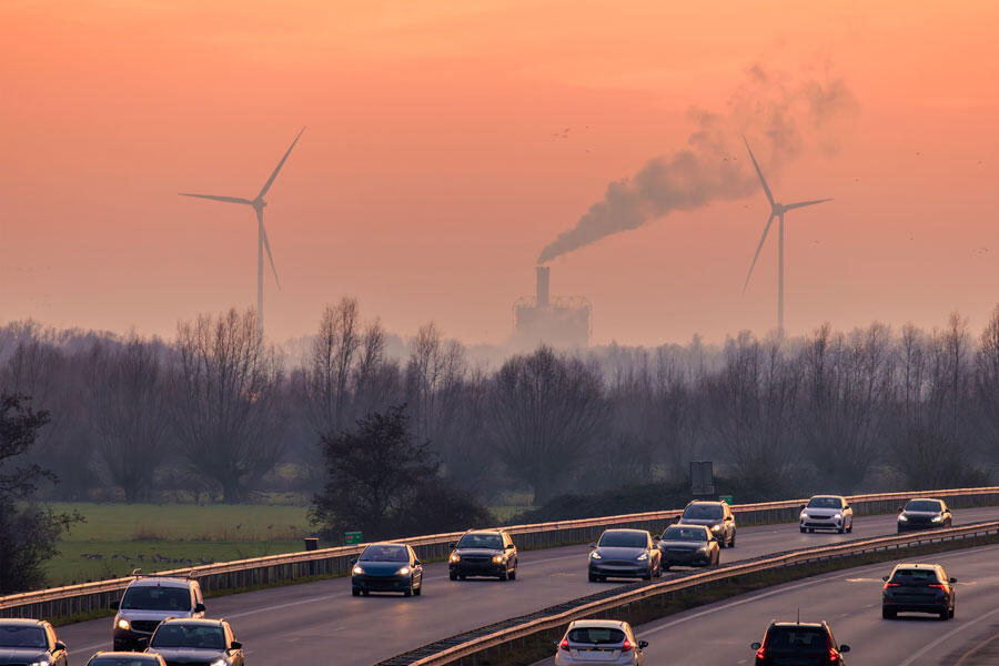 Voitures sur une autoroute en Europe, et en arrière-plan, des fumées d’usine et des éoliennes