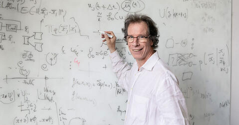 Portrait de Stéphane Mallat devant un tableau blanc couvert de formules mathématiques