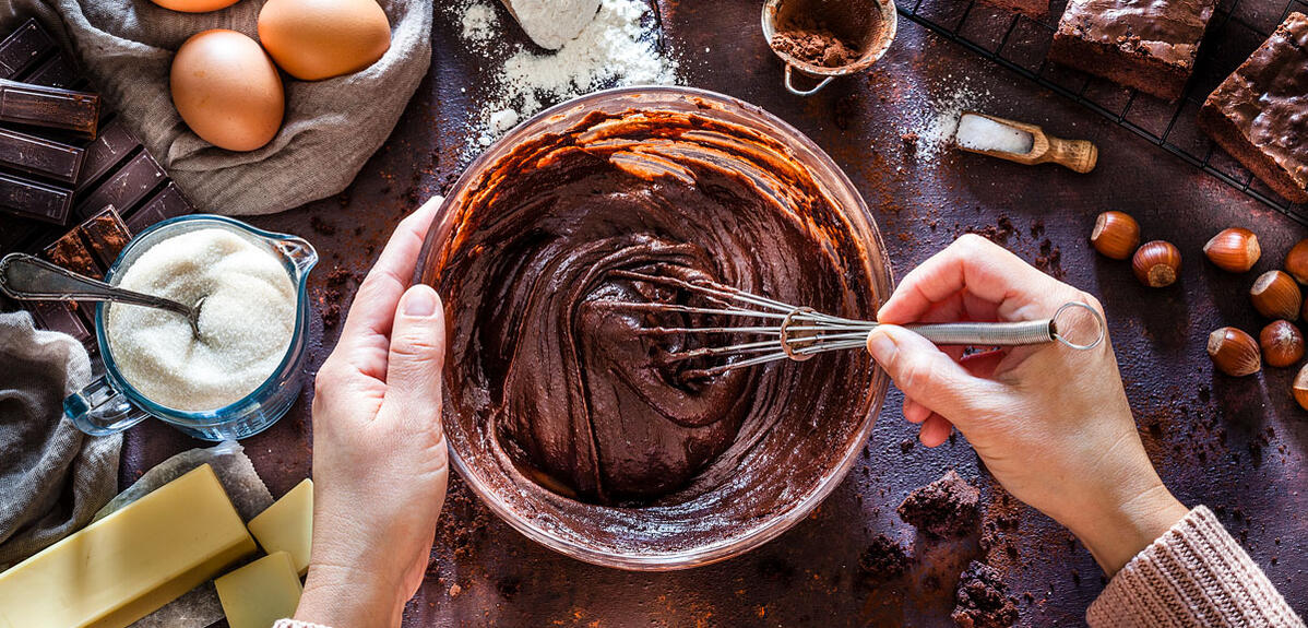 Des mains mélangent une pate chocolatée avec un batteur, les ingrédients du gâteau sont disposés autour 
