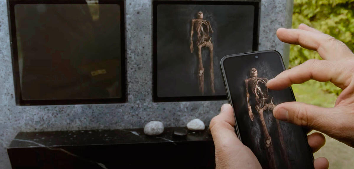©Prospero Pictures / SBS Productions / Saint Laurent - Collection ChristopheL - Film de David Cronenberg 2024 image du film où une main touche un téléphone qui montre l’intérieur de la tombe et on voit le squelette