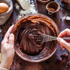 Des mains mélangent une pate chocolatée avec un batteur, les ingrédients du gâteau sont disposés autour 