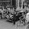 Dans une rue de Paris en 1940, un agent de police retient derrière une ligne de clous divers engins roulants